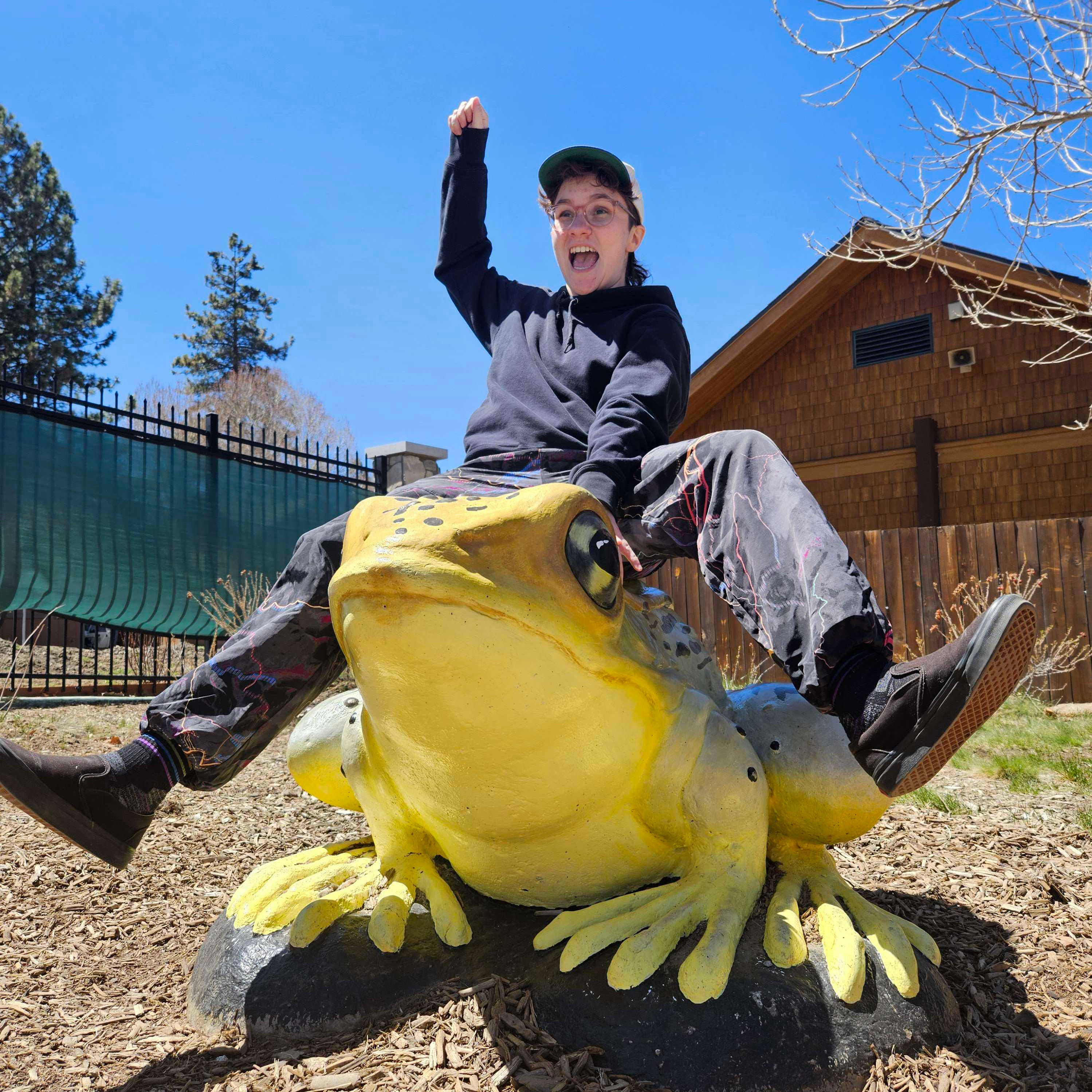 a cropped photo of alex pretending to ride a frog statue at a zoo. they have on a black hoodie, a snapback hat, patterened colorful pants, and black sneakers. they have their hand up as if they are a rodeo cowboy.