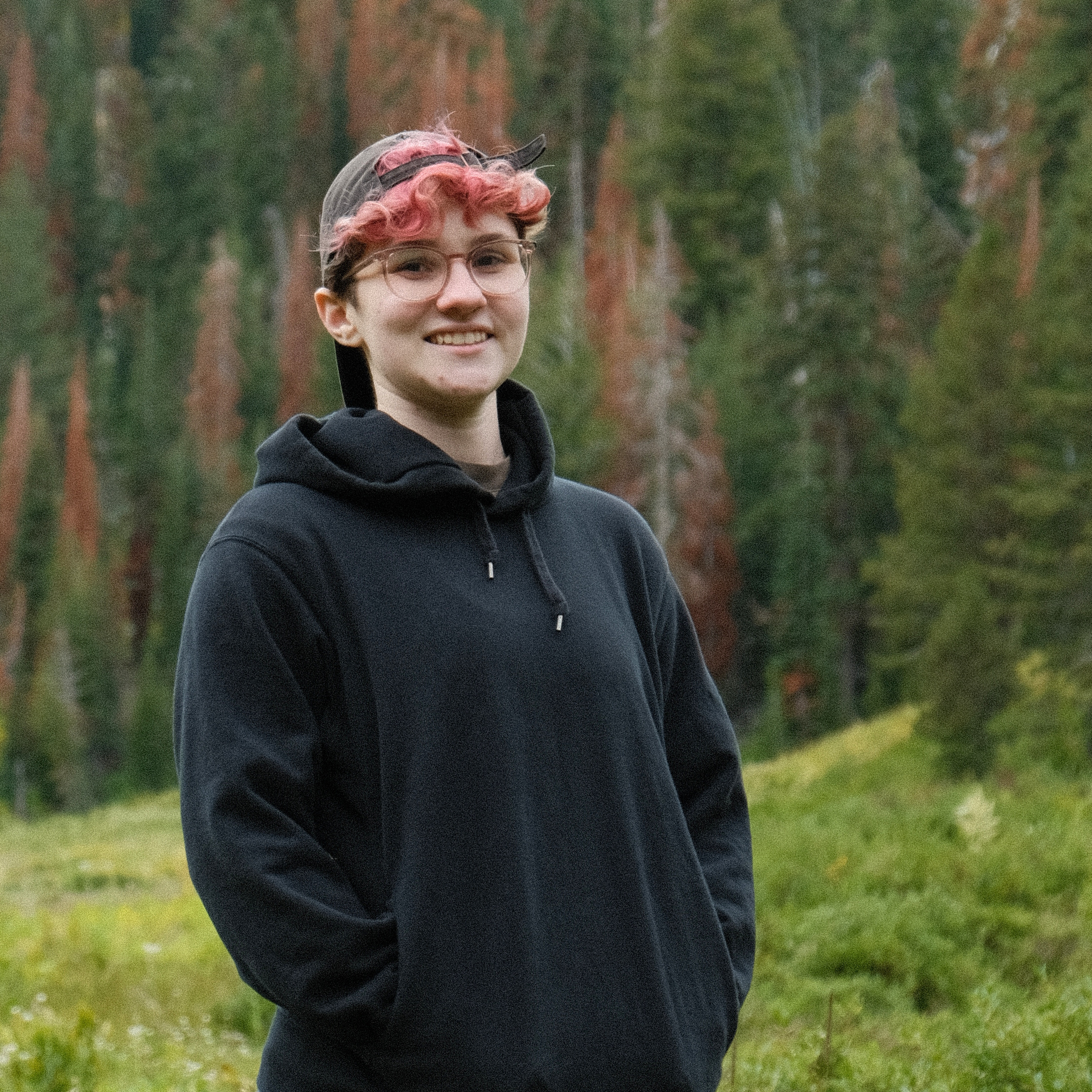 a cropped photo of alex sitting on a bench in the woods. they have on a red tank top, black shorts, a black hat, pink clear glasses, and a backpack. they are looking away from the camera and have their hands clasped together with their elbows rested on their knees.